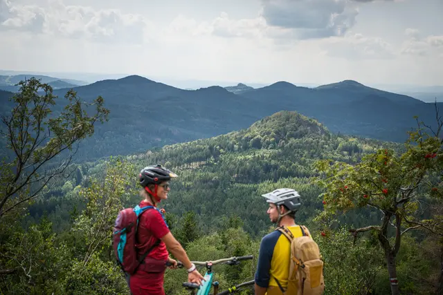 Zwei Mountainbiker blicken von einem Felsen über die bewaldete Berglandschaft.