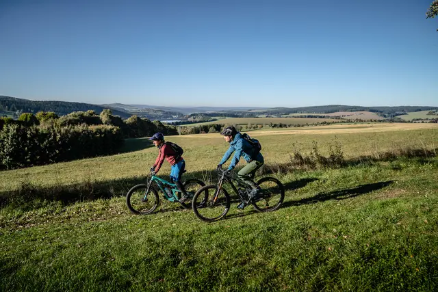Zwei Mountainbiker fahren nebeneinander durch eine sonnige Mittelgebirgslandschaft.