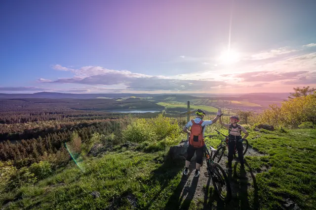 Zwei Mountainbiker heben ihre Räder vor einem weiten Sonnenuntergangspanorama an.