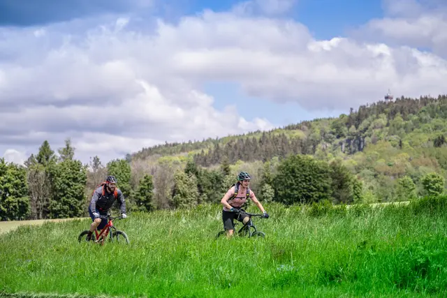 Zwei Mountainbiker fahren über eine Wiese vor bewaldeten Mittelgebirgshängen.
