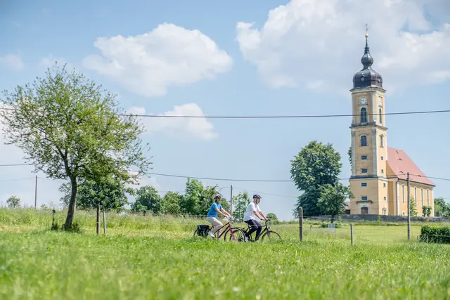 Zwei Radgäste fahren über eine Wiese an einer Dorfkirche vorbei.