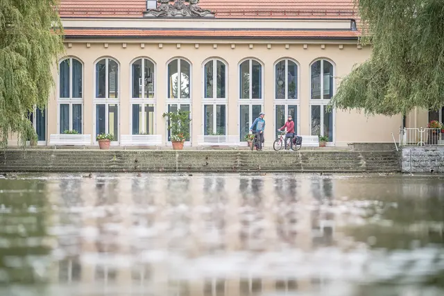 Zwei Radgäste fahren an einem historischen Gebäude mit Wasserbecken entlang.
