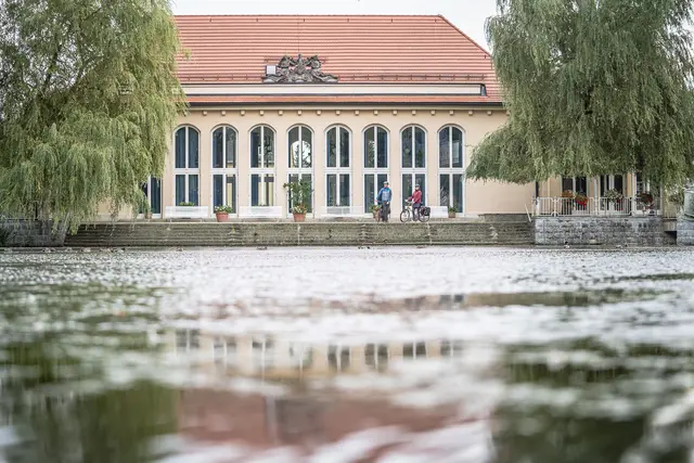 Zwei Radgäste passieren ein historisches Gebäude an einem Wasserbecken.