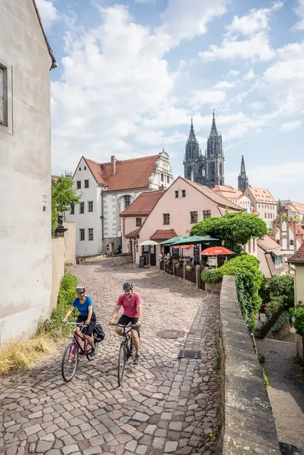 Zwei Radgäste schieben ihre Fahrräder durch eine historische Gasse mit Blick auf den Meißner Dom.