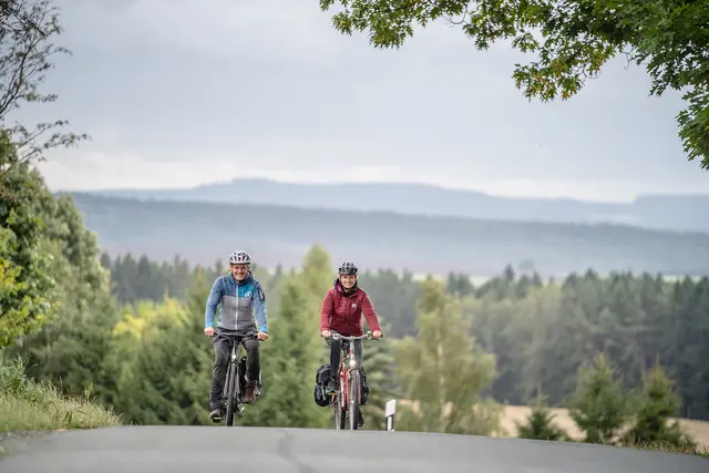 Zwei Radgäste fahren auf einer Straße vor einer bewaldeten Mittelgebirgslandschaft.