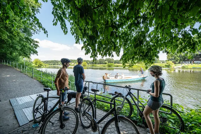 Radgäste stehen mit ihren Fahrrädern am Ufer eines Badesees.