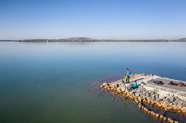 Radgäste sitzen mit ihren Fahrrädern auf einer Mole in einem großen See.