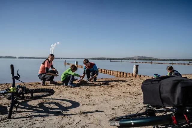 Eine Familie sitzt während ihrer Radtour am Ufer eines Sees; die Fahrräder liegen im Vordergrund.