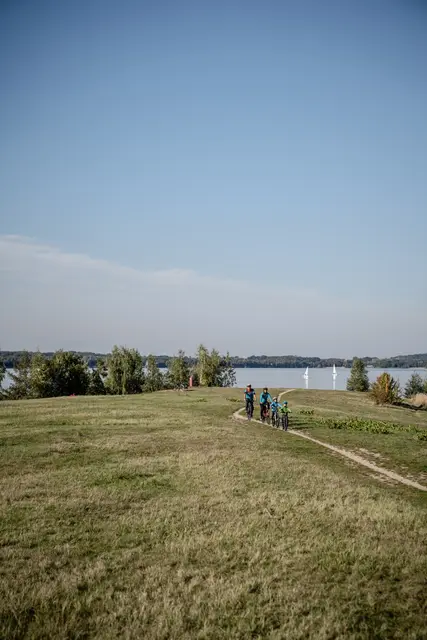 Ein schmaler Radweg führt durch eine offene Landschaft zu einem See.